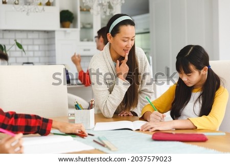 Asian mother helping daughter with school work, with brother in foreground and father in background. busy family working together at home in their kitchen.