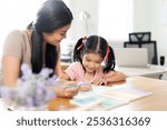 Asian mother helping her daughter with homework, sitting in living room interior, copy space. 