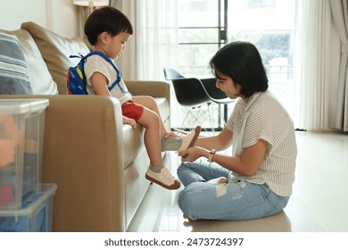 Asian Mother helps young son put on shoes in living room. Boy with backpack sits on couch while mother ties shoelaces. Morning routine, parent-child bonding, preparation for school. - Powered by Shutterstock