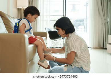 Asian Mother helps young son put on shoes in living room. Boy with backpack sits on couch while mother ties shoelaces. Morning routine, parent-child bonding, preparation for school. - Powered by Shutterstock