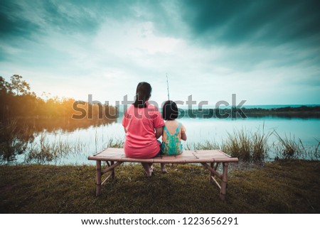 Asian mother and her daughter sit on the banks of a tranquil evening lagoon.The girl is holding a fishing rod.
