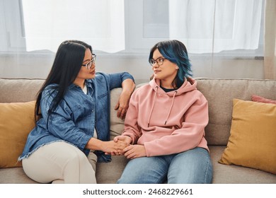 An Asian mother and her teenage daughter in casual wear engage in a deep conversation while sitting on a cozy couch. - Powered by Shutterstock