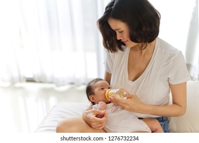 Asian mother holding and feeding baby from milk bottle. Portrait of cute newborn baby being fed by her mother using bottle on sofa. Loving woman giving to drink milk to her baby.