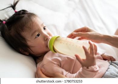 Asian Mother Holding And Feeding Baby From Milk Bottle. Portrait Of Cute Newborn Child Being Fed By Mother On Bed In Warm Bedroom. Loving Woman Giving Nutrition Food To Kid Drinking Mom Milk At House.