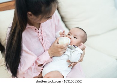 Asian Mother Holding And Feeding Baby From Milk Bottle. Portrait Of Cute Newborn Child Being Fed By Mother On Sofa In Living Room. Mom Giving Nutrition Food To Kid Drinking The Milk At House.