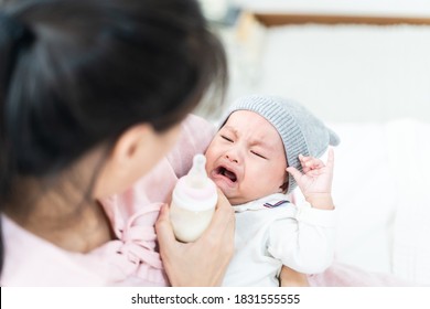 Asian mother holding and feeding crying baby from milk bottle. Portrait of cute newborn child being fed by mother on sofa in living room. Mom giving nutrition food to kid drinking the milk at house.