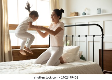 Asian Mother Holding Hands Of Little Toddler Daughter While She Jumping On Bed. Family Spend Together Active Energetic Funny Time In The Morning In Bedroom. Happy Motherhood, Cozy Modern Home Concept