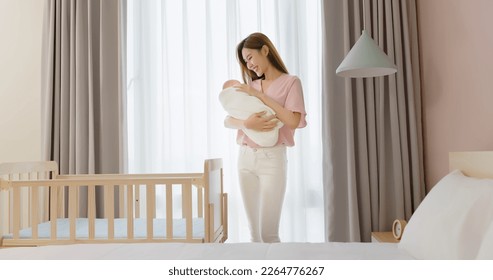 asian mother is hugging comforting cute newborn baby to sleep and standing besides crib at home