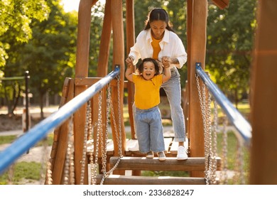 Asian Mother With Infant Daughter Walking Swinging Bridge Together, Child Holding Mom's Hands Enjoying Fun Outside On Playground In City Park. Outdoor Entertainment for Toddler Baby - Powered by Shutterstock