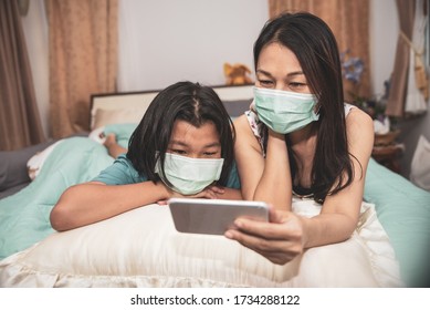 Asian Mother And Kid Wear Mask And Using Mobile Phone Together. Mother And Daughter Lie On The Bed And Learn Online Study At Home. Happy Family In New Normal Lifestyle Concept