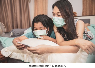 Asian Mother And Kid Wear Mask And Using Mobile Phone Together. Mother And Daughter Lie On The Bed And Learn Online Study At Home. Happy Family In New Normal Lifestyle Concept.