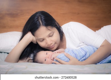 Asian Mother Lays On Her Bed In Her Bedroom With Her Freshly Bathed Mixed Race 10 Day Old Newborn Baby Boy Wrapped In A Clean Blue Towel