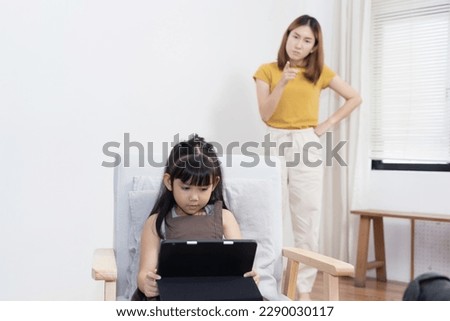 Asian mother looking at her daughter playing game on tablet while studying