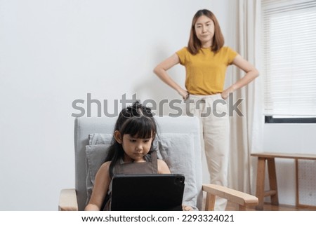 Asian mother looking at her daughter playing game on tablet while studying