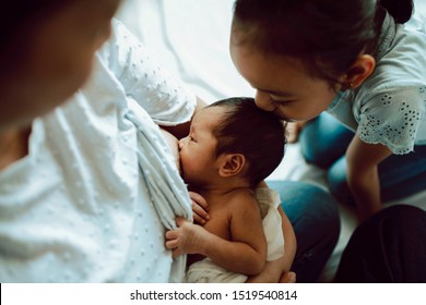 Asian Mother Nursing Newborn. Mom Holding Her Child While Breastfeeding. New Mommy Holds Baby While Sister Kisses Brother’s Head . Woman And New Born Boy On The Bed At Home. Family Bonding Time.