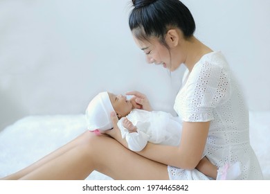 An Asian Mother Play With Her Baby Laying Down On Her Lift Knee On White Bed With Happiness And Both Smile, Bonding Of Dauther And Mom, Cheerful, Lovely Moment. Nusing, Caring Concept.