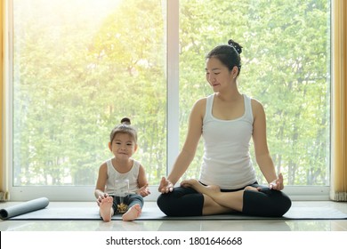 Asian Mother Practiced Yoga By Sitting In Lotus Position At Home With A Daughter Sitting Beside And Enjoy Eating Snack With Smiling Face While Workout At Home. Mom And Kid Doing Activity At Home 