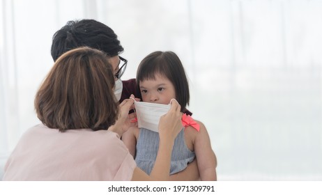 Asian Mother Putting Medical Face Mask For Young Disabled Child, Down Syndrome Or Trisomy 21 Daughter Sitting On Father's Lap During Pandemic Outbreak Of Coronavirus Or Covid 19.