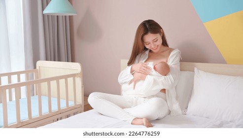 asian mother sitting on bed is breastfeeding her baby - holding and looking infant