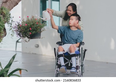 Asian Mother With Smiling Face As She Pushes Her Son's Wheelchair Water The Plants In The Garden, Family Is Support To Develop Skills For Life And Happy, Mental Health And Mother Day A Concept.