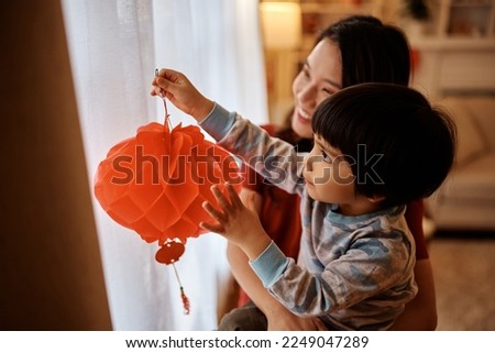 Asian mother and son hanging red lanterns on window while decorating home for Chinese New Year.