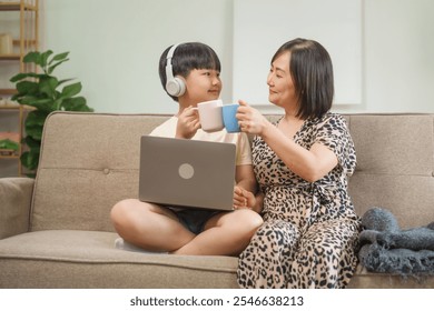 An Asian mother and son sit at the table and sofa, sharing time together while looking at a computer, tablet, and phone. They bond through technology and family activities - Powered by Shutterstock