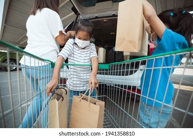 Asian mother and two daughters wearing protection mask helping to put stuffs in the shopping trolley into trunk of car together in parking during coronavirus pandemic as new normal lifestyle. - Shutterstock ID 1941358810