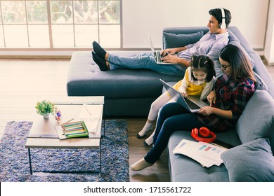 Asian Mother Using Laptop Computer To Work On Sofa In Living Room While Teach And Explain Her Half Little Cute Daughter. Caucasian Father Sitting And Working With Notebook In Background, Happy Family.