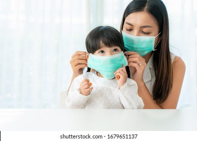 Asian Mother Wearing Wearing To Her Daughter Healthy Face Mask Sitting In Living Room At Home To Prevent PM2.5 Dust, Smog, Air Pollution And COVID-19. Healthcare Concept.