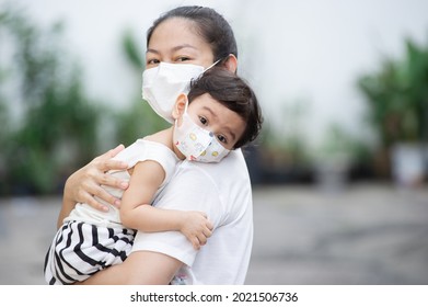 Asian mother wearing white face mask holding her little toddler baby girl up close who is her daughter wear baby face mask, look at camera, COVID-19 concept