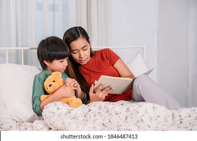 Asian Mother Work Home Together With Son. Mom And Kid Reading Fairy Tale Before Sleep On The Bed. Woman Lifestyle And Family Activity.