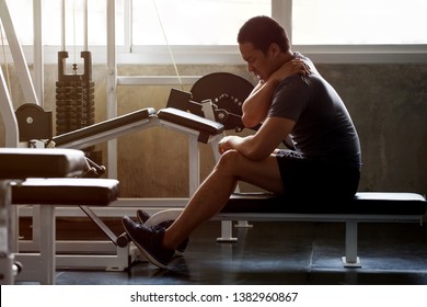 Asian Muscular Man Suffering From Shoulder Injury While Exercising At Gym In Morning Light . Workout , Exercises , Bodybuilder Training , Sport Guy Rubbing His Back And Neck Pain .  Backache