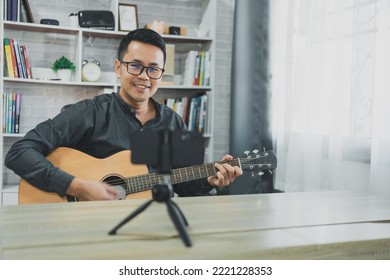 Asian Music Teacher Man Broadcasting Live Guitar Lessons To Students Online Using Mobile. Asian Male Musician Playing Guitar Singing Live Video Conferencing At Home. Live Concert Music From Anywhere.