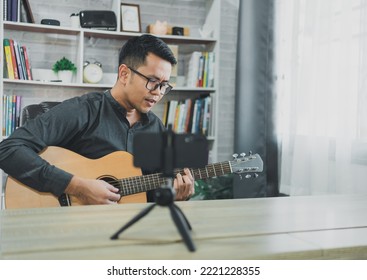 Asian Music Teacher Man Broadcasting Live Guitar Lessons To Students Online Using Mobile. Asian Male Musician Playing Guitar Singing Live Video Conferencing At Home. Live Concert Music From Anywhere.