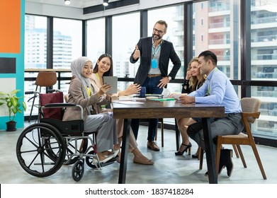 Asian Muslim Business Woman In Hijab Headscarf Sitting On Wheelchair Presenting Of Her Work To Corporate Colleagues In Meeting In The Modern Office. Diverse Corporate Colleagues And Multicultural