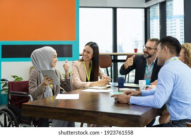 Asian Muslim Business Woman In Hijab Headscarf Sitting On Wheelchair Presenting Of Her Work To Corporate Colleagues In Meeting In The Modern Office. Diverse Corporate Colleagues And Multicultural