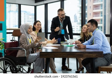 Asian Muslim Business Woman In Hijab Headscarf Sitting On Wheelchair Presenting Of Her Work To Corporate Colleagues In Meeting In The Modern Office. Diverse Corporate Colleagues And Multicultural