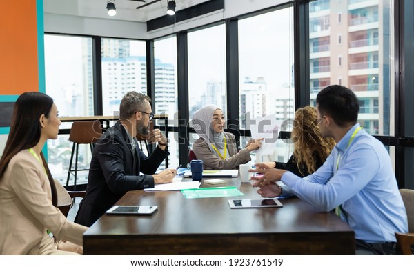 Asian Muslim business woman in hijab\
headscarf sitting on wheelchair presenting of her work to corporate\
colleagues in meeting in the modern office. diverse corporate\
colleagues and\
multicultural