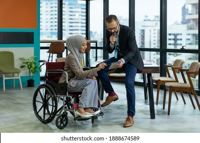 Asian Muslim businesswoman sitting on wheelchair presenting business graph on paper to manager. Smiling group of diverse corporate colleagues in the modern office. Diversity or multicultural in office