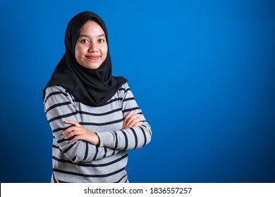 Asian muslim college student girl wearing hijab smiling friendly with arms crossed against blue background
