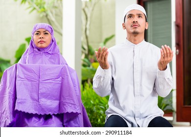 Asian Muslim Couple, Man And Woman, Praying At Home Sitting On Prayer Carpet In Their House In Front Of The Tropical Garden