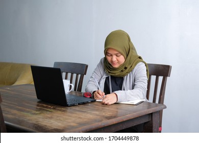 An Asian Muslim Female Work Remotely From Home With Laptop And Notes At Wooden Dining Table Due To Quarantine. Video Conference With Colleague To Discuss Work.