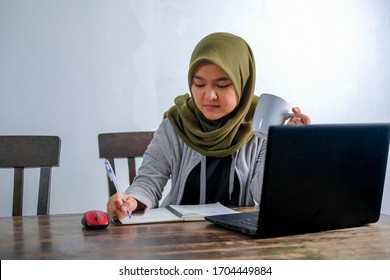 An Asian Muslim Female Work Remotely From Home With Laptop And Notes At Wooden Dining Table Due To Quarantine. Video Conference With Colleague To Discuss Work.