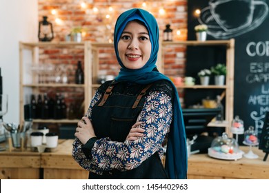 Asian Muslim Girl Wearing Hijab Working As Waitress In Cafe Bar. Young Elegant Islam Lady Crossed Arms Small Start Up Business Owner In Coffee Shop. Arabic Woman Staff Face Camera Smiling Attractive