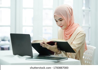 Asian Muslim High School Student Girl Reading The Book While Doing A Educational Project  In Front Of Laptop Screen. Learning, Studying, Technology And Education Concept.                       