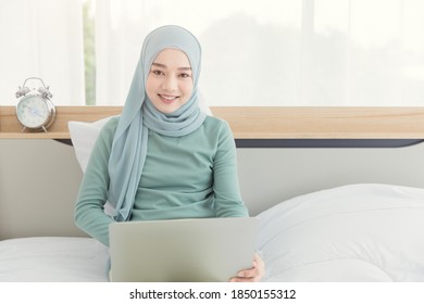 Asian Muslim Islamic Woman With Hijab Happy Enjoy Online Remote Work At Home With Her Laptop On The Bed.