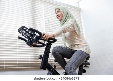 Asian muslim islamic women cycle at home using stationary exercise bike, healthy lifestyle workout with sport bike exercises, daily leisure activity routine, happy smiling with enjoyment. - Powered by Shutterstock