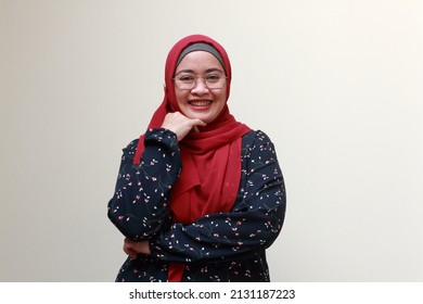 An Asian Muslim Lady Standing With One Hand On Her Chin And Smiling.