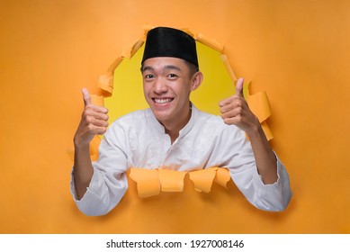 Asian muslim man smiling and showing thumbs up poses through torn yellow paper hole, wearing muslim cloth with skull cap.