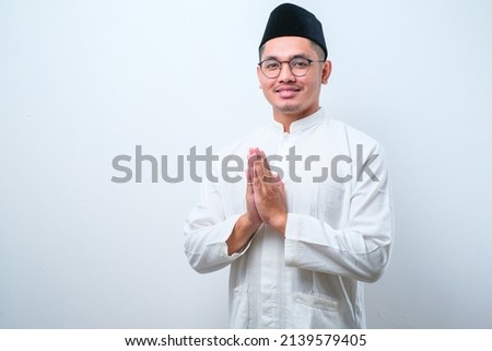 Asian Muslim man wearing glasses smiling to give greeting during Ramadan and Eid Al Fitr celebration over white background